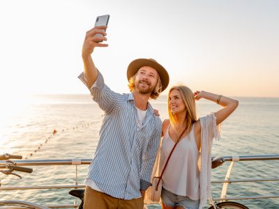young attractive smiling happy man and woman traveling on bicycles taking selfie photo on phone camera, romantic couple by the sea on sunset, boho hipster style outfit, friends having fun together